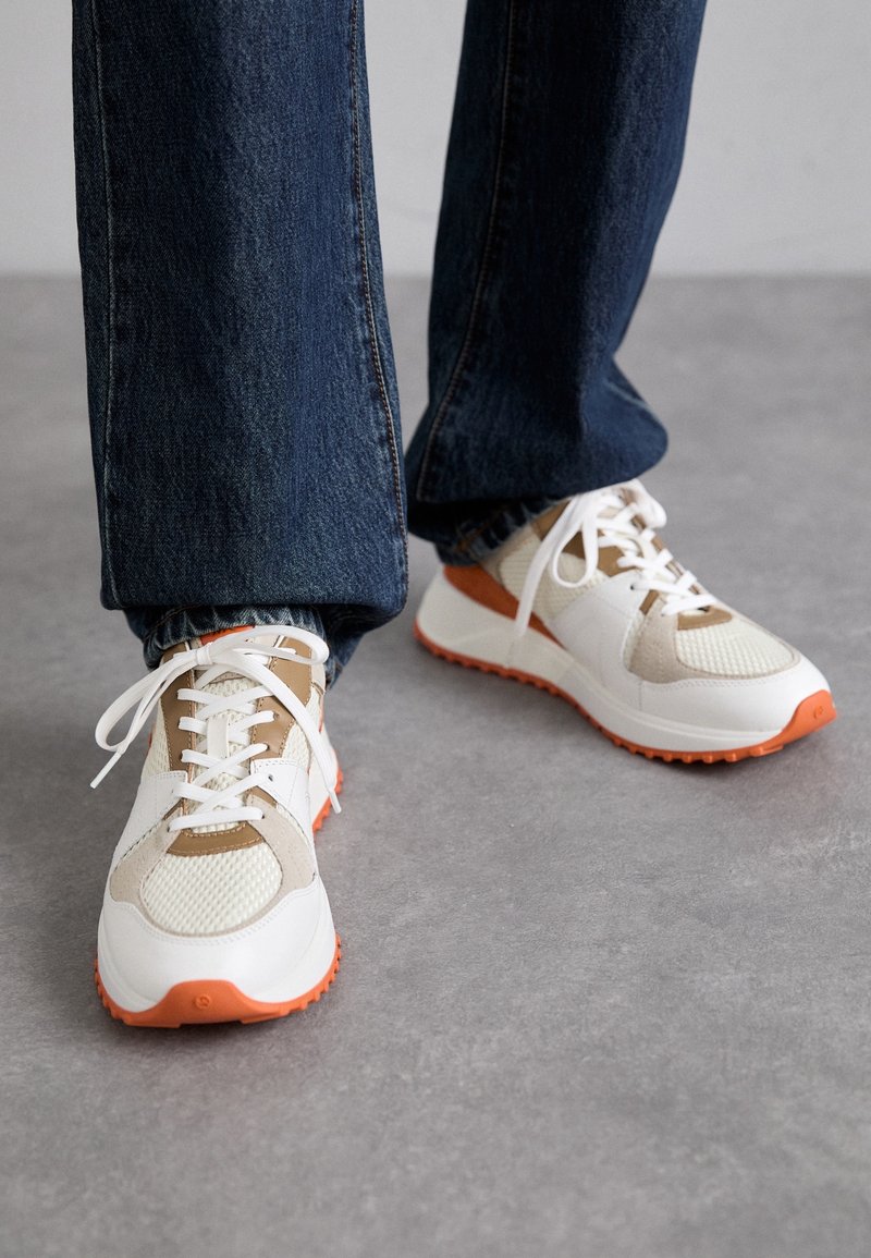 Personne portant des baskets blanches et beige avec des semelles orange et un jean bleu foncé, debout sur un sol gris.