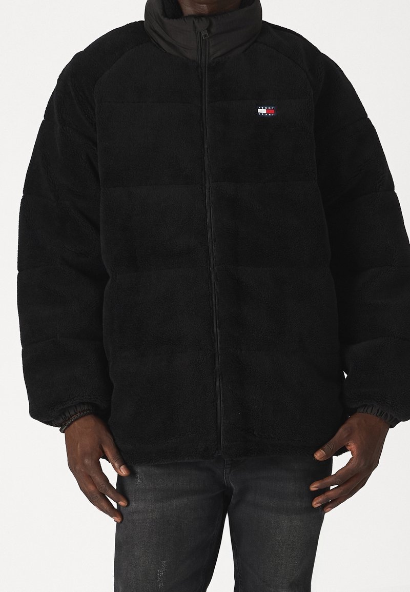 Homme portant une veste polaire noire zippée avec un petit logo rectangulaire sur la poitrine gauche, associée à un jean foncé.