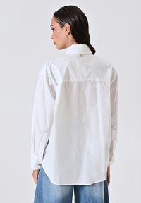 Camicia bianca con bottoni, maniche lunghe, orlo arrotondato e dettagli del logo sul retro. Realizzata in tessuto liscio, abbinata a pantaloni in denim.