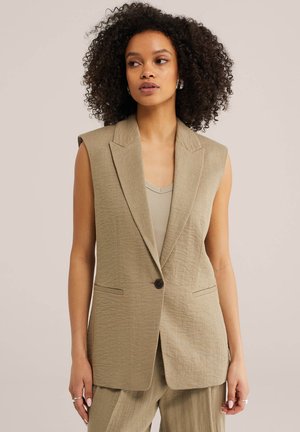 Frau mit lockigem Haar trägt ein ärmelloses beiges Blazer über einem Tank-Top und passenden weit geschnittenen Hosen, stehend vor einem neutralen Hintergrund.