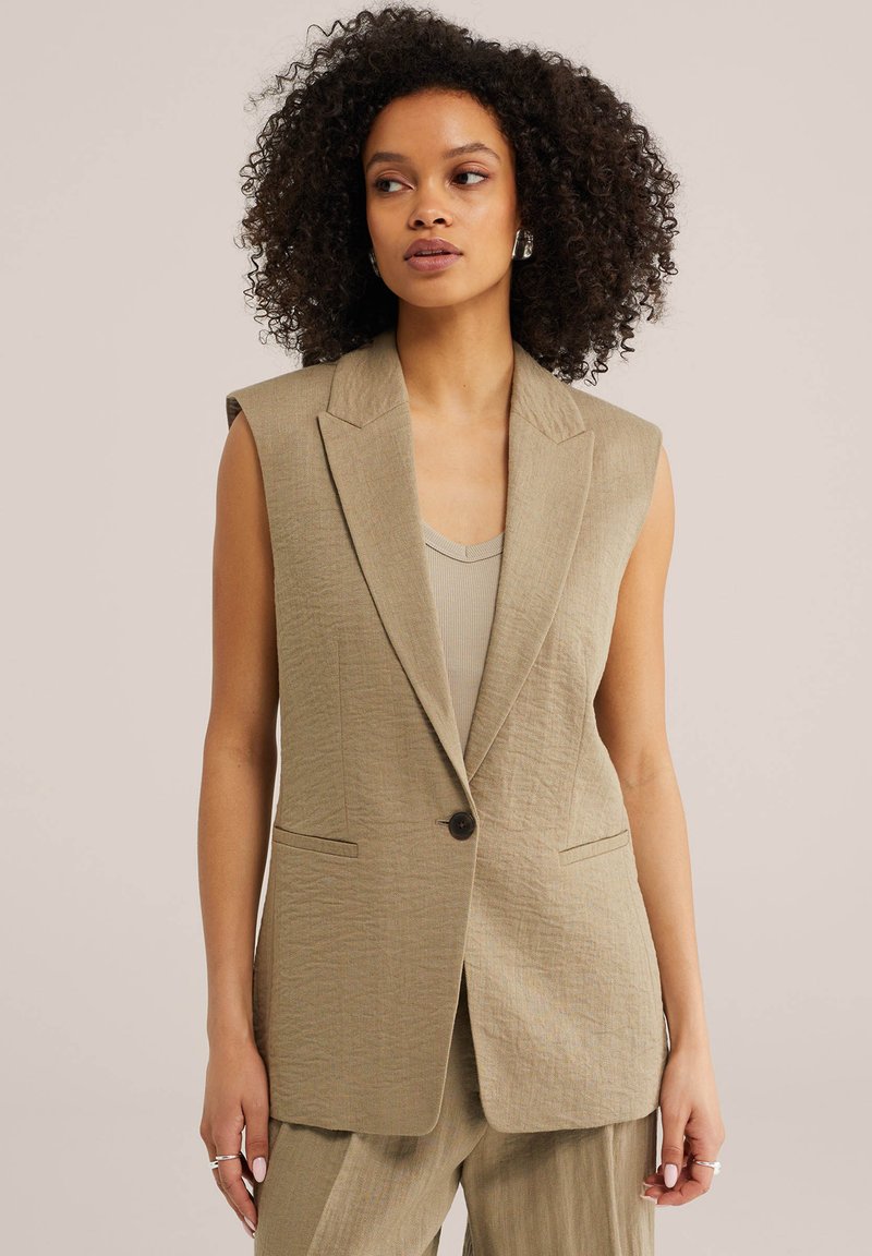 Žena s kučeravými vlasmi má na sebe béžový bezrukávový blejzer cez tielko a zodpovedajúce nohavice širokého strihu, stojaca na neutrálnom pozadí.