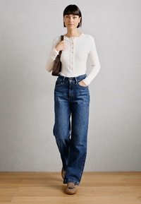 White ribbed long-sleeve top with button detail, paired with high-waisted blue wide-leg jeans. Brown handbag and light-colored shoes.