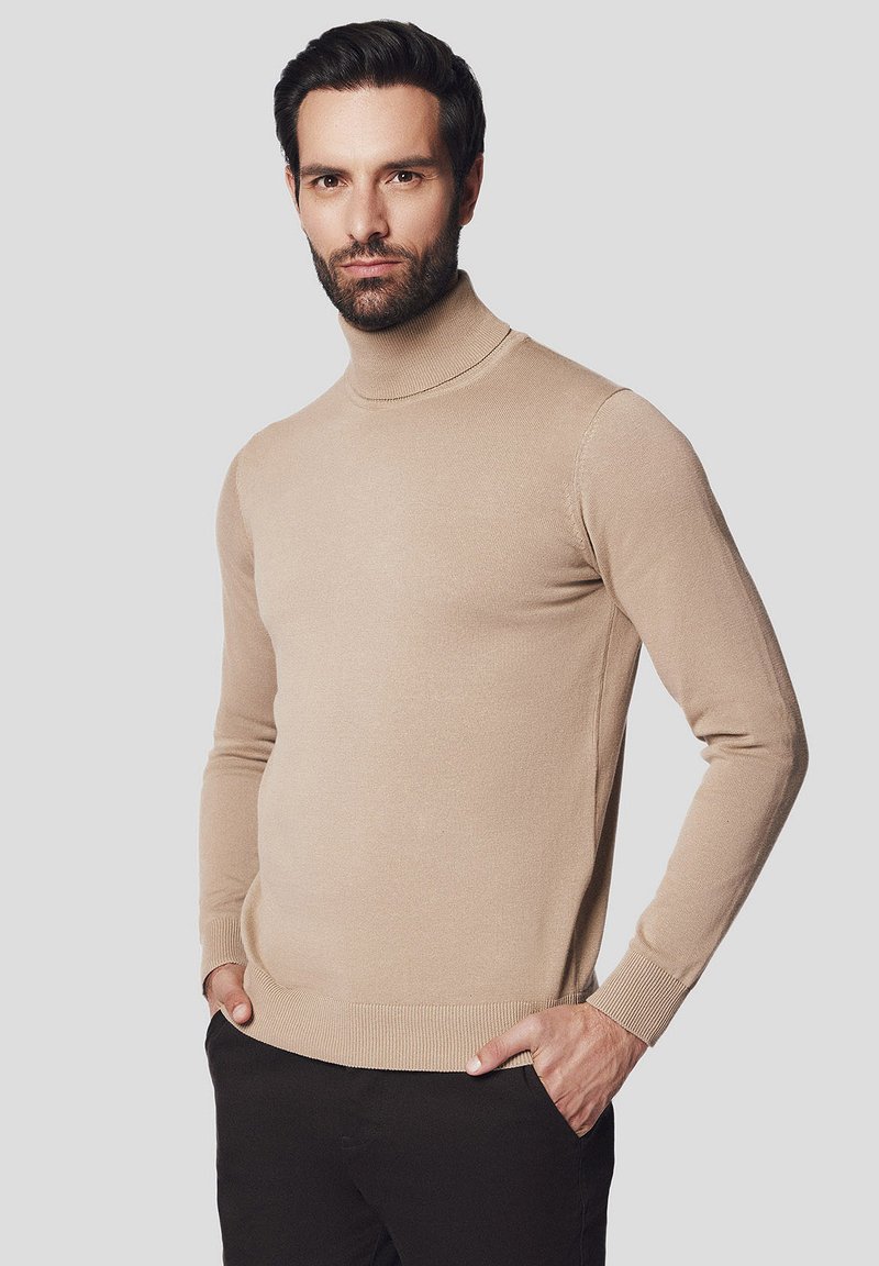 Beżowy sweter z golfem o dopasowanym kroju, z prążkowanymi mankietami i dołem, wykonany z miękkiej dzianiny, noszony na ciemnych spodniach.