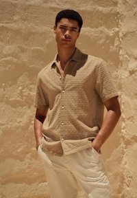 Une chemise à manches courtes, de couleur brun clair, avec un motif texturé et des boutons, associée à un pantalon beige clair sur un fond neutre.