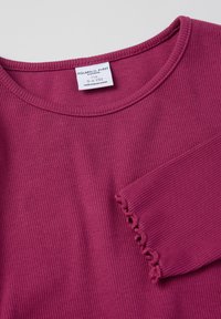 Violetti ribbattu pitkähihaine paita, jossa on pyöreä kaula-aukko ja tasku, jossa on aaltoileva reuna. Hoito-ohjeet näkyvät kaula-aukon sisäpuolella.