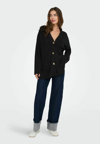 Svart oversized cardigan med V-ringning, prydd med guldknappar, två framfickor och en texturerad yta, i kombination med vida jeans.