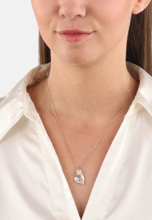 Collar de oro rosado con un colgante en forma de corazón que presenta un acabado iridiscente y pequeños adornos florales. Llevado sobre una camisa de satén blanca.