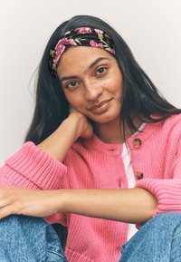 Pink gestrickte Strickjacke mit rundem Ausschnitt und Holzknöpfen. Schwarzes Blumen-Stirnband. Lange dunkle Haare, helle Haut mit Sommersprossen.