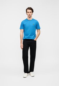 Μπλε βαμβακερό T-shirt με στρογγυλή λαιμόκοψη, συνδυασμένο με μαύρο παντελόνι με ίσια γραμμή και λευκά αθλητικά παπούτσια. Διακρίνεται κεντητό λογότυπο στο στήθος.