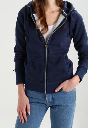 Kobieta ubrana w granatową bluzę z zamkiem na suwak na białą koszulkę, z jedną ręką w kieszeni, w niebieskich dżinsach, na jednolitym tle.