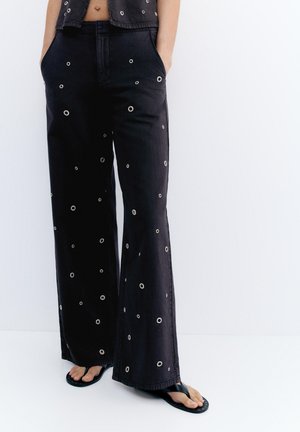 Zwarte wijde broek met witte ronde eyeletpatronen, gedragen met zwarte teenslippers, handen in de zakken tegen een effen witte achtergrond.