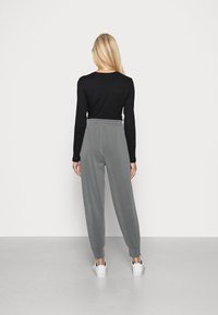 Blond kvinna som står vänd mot en vit vägg, klädd i en svart långärmad tröja, grå högmidjade byxor och vita sneakers.
