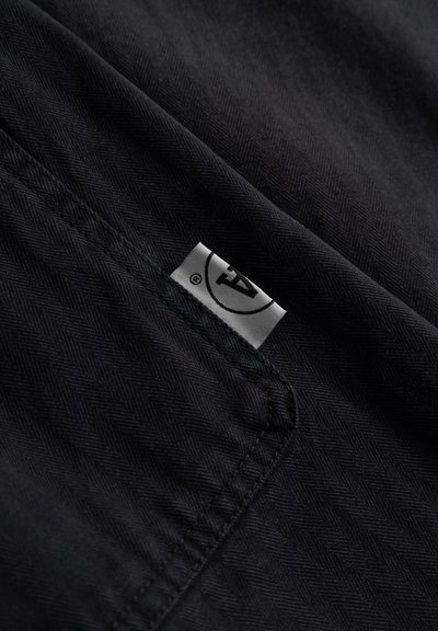 Tissu noir avec un motif en chevrons ; comporte une petite étiquette blanche avec un logo ; texture lisse ; plié et partiellement visible.