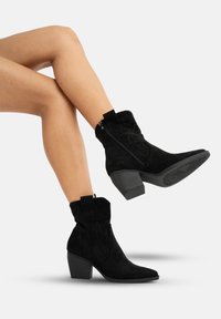 Botas de ante negras hasta el tobillo con un diseño cosido, puntera puntiaguda y un tacón grueso. Presentan una cremallera en el lateral y una textura suave.