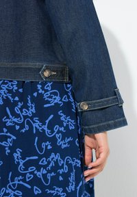 Tmavě modrá džínová bunda se stříbrnými knoflíky, kombinovaná s modrými šaty s ručně psanými milostnými frázemi v světlejší modré barvě.