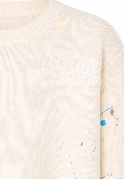 Maglione lavorato a maglia color crema con numeri bianchi da 0 a 22 e numero 6 cerchiato sul petto, con schizzi di vernice blu e grigia sulla manica destra e sul corpo.