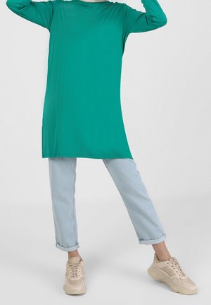 Personne portant une tunique manches longues de couleur turquoise, un jean bleu clair retroussé et des baskets beiges, debout les jambes écartées sur un fond uni.