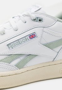 Vita lädersneakers med ljusgröna accenter och en texturerad off-white gummisula. Har Reebok-logotyp och brittisk flagga.