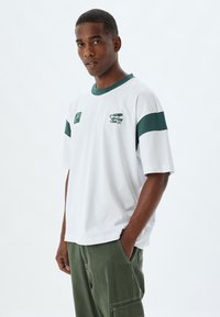 Camiseta blanca de algodón con detalles en verde. Presenta un cuello redondo acanalado, mangas cortas y un pequeño logo en el pecho. Diseño de corte holgado.