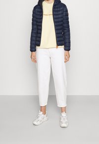 Marineblå pufferjakke med hætte, lynlåslukning, orange detaljer, gul t-shirt med trykt tekst, hvide ankellange bukser og lyse sneakers.