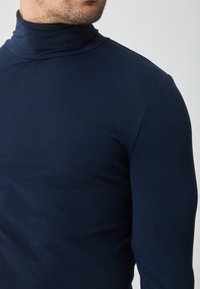 Tricou cu guler înalt de culoare bleumarin, fabricat dintr-un material neted și elastic. Are un design ajustat, cu mâneci lungi și un guler înalt.