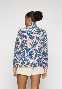 Fleecejacke mit hohem Kragen, die ein mehrfarbiges, abstraktes Blumenmuster auf einem hellen Hintergrund und kontrastierenden gelben Akzenten zeigt.