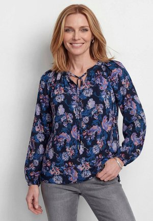 Frau mit schulterlangen blonden Haaren, die eine langärmelige marineblaue Bluse mit lila und rosa Blumenmustern und graue Hose trägt, lächelt.