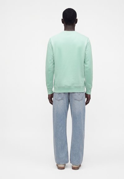 Homme portant un sweat-shirt vert clair à manches longues et un jean bleu clair ample, debout de dos sur un fond blanc uni.