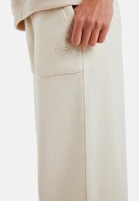 Beige sweatpants gemaakt van zachte stof met een gemêleerde afwerking. Bevat een zijzak en een reliëflogo op de voorkant.