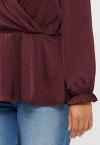 Burgundy blouse with a wrapped design and ruffled cuffs, paired with blue jeans, highlighting the fabric's texture and fit.