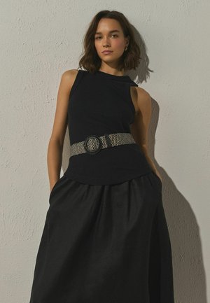 Junge Frau mit schulterlangem Haar, die ein ärmelloses schwarzes Oberteil, einen breiten gewebten Gürtel und einen schwarzen Rock trägt und vor einer strukturierten Wand steht.