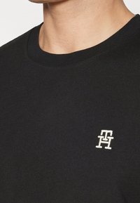 Černé bavlněné tričko s kulatým výstřihem, na hrudi má bílé vyšívané logo. Hladká textura, ležérní design.