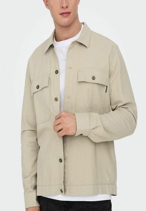 Chaqueta de botonadura ligera color beige, con dos bolsillos en el pecho con botones, un cuello recto y mangas largas. Llevada sobre una camiseta blanca.