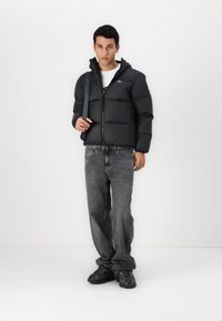 Veste matelassée noire avec capuche, fermeture éclair à l'avant et texture matelassée. Assortie avec un jean en denim gris et des chaussures de sport noires.