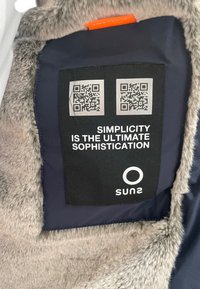 Fodera di giacca blu navy con pelliccia sintetica, che presenta un'etichetta per la cura del tessuto nera. L'etichetta ha due codici QR e il testo: "La semplicità è la massima sofisticazione."