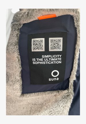 Fodera di giacca blu navy con pelliccia sintetica, che presenta un'etichetta per la cura del tessuto nera. L'etichetta ha due codici QR e il testo: "La semplicità è la massima sofisticazione."