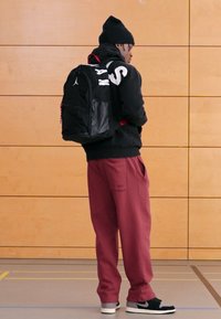 Fekete hátizsák piros díszítéssel, szürke melegítőnadrág és fekete sportcipő. A háttérben fa falburkolat és a tornaterem padlóvonalai láthatók.