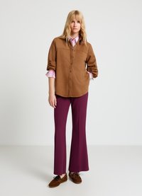 Chemise à boutons en daim marron portée sur une chemise à carreaux rose et blanc, associée à un pantalon évasé prune et des chaussures style mocassins marron.