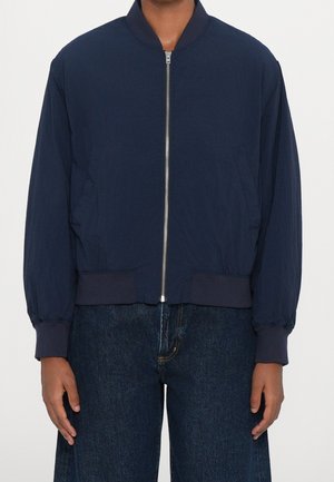 Personne portant une veste bomber bleu marine avec une fermeture éclair frontale et un jean bleu foncé, debout devant un fond blanc uni.