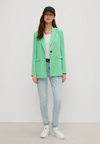 Grüne Blazer mit einem einzelnen Knopf, kombiniert mit einem weißen T-Shirt und hellblauen Jeans. Eine schwarze Mütze und weiße Sneakers runden das Outfit ab.