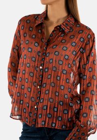 Blusa roja transparente con un patrón geométrico en azul marino y blanco. Presenta un escote con cuello y cierre de botones en la parte delantera.