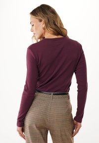 Frau mit welligem blonden Haar, die goldene Creolen trägt, ein enges, geripptes lila Langarmoberteil und karierte, hoch taillierte Hosen mit einem schwarzen Gürtel.