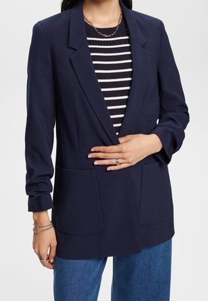 Mujer con blazer azul marino sobre suéter de rayas blancas y negras y vaqueros azules, con anillos plateados y pulseras de cadena.