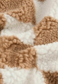 Gros plan sur un tissu en polaire doux et texturé avec un motif en patchwork brun et blanc, montrant sa surface duveteuse et confortable.