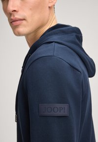 Marineblauer Hoodie mit Kordelzug, glatter Textur und einem gummierten schwarzen Logo-Patch am linken Arm. Mit einem geteilten Design an der Schulter.