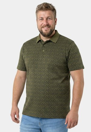 Smilende mand iført en grøn mønstret poloshirt og lyseblå jeans, stående mod en ensfarvet, lys grå baggrund.