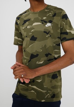 Mężczyzna w zielono-czarnej koszulce Nike moro, stoi ze złączonymi rękami z przodu, na tle jednolitego jasnoszarego tła.