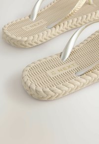 Beige Flip-Flops mit gewebten strukturierten Sohlen, ausgestattet mit metallischen Riemen und einem dezenten Markenlogo auf der Fußbett.