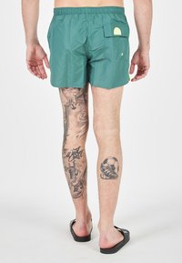 Pantaloni da bagno verdi con vita elasticizzata, dotati di una tasca posteriore e dettagli con logo. Tatuaggi visibili sulle gambe includono design sportivi e artistici.