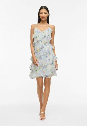 Femme portant une robe sans manches à fleurs pastel avec des volants superposés et des bretelles fines, debout jambes croisées et portant des talons argentés à fines lanières.